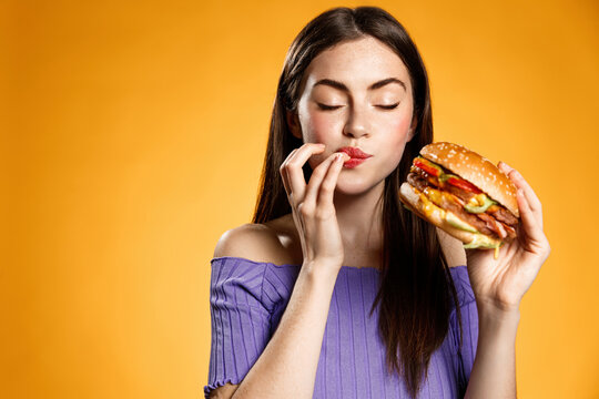 Woman Eating Cheeseburger With Satisfaction. Girl Enjoys Tasty Hamburger Takeaway, Licking Fingers Delicious Bite Of Burger, Order Fastfood Delivery While Hungry, Standing Over Orange Background