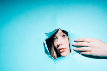 Young girl face with makeup looking to the side through a hole in a blue background revealing it hand