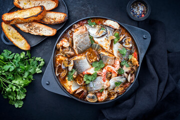 Modern style traditional Spanish seafood zarzuela de pescado with fish, king prawns and venus clams served with barbecued bread in red sauce as top view in design pot
