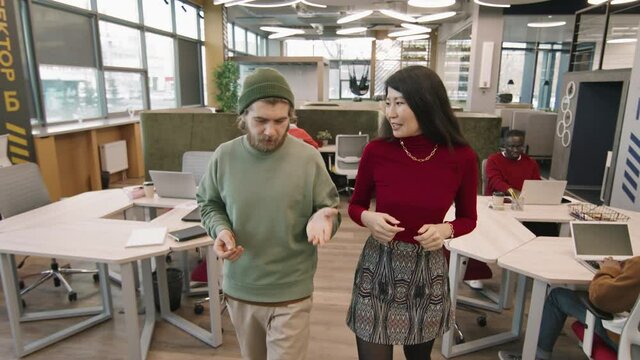 Tracking slowmo of young Asian businesswoman and bearded businessman in beanie hat walking in open-plan office and talking