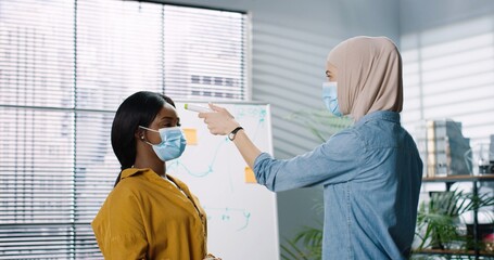 Young woman in protective mask measures the temperature of colleagues who come to office using electronic thermometer. Concept of new rules of safety and social distance.