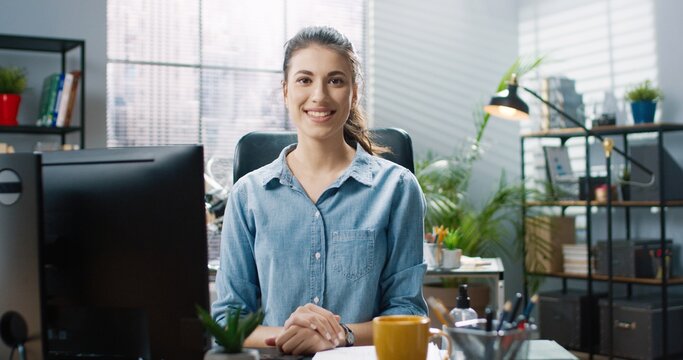 Portrait of young beautiful girl in blue shirt sitting in office at table and takes off protective medical mask smiling looking at camera.