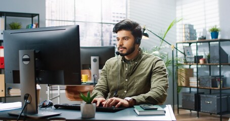 Portrait of joyful handsome young Hindu male worker in headset sitting at table in modern cabinet...
