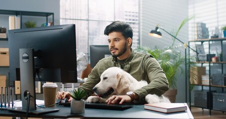 Young handsome Hindu guy sits in office with his big Labrador Retriever dog and works on computer completing task.