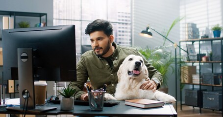 Portrait of joyful Hindu handsome man sitting in cabinet at work with his cute dog pet in good mood typing on computer browsing online. Positive emotions, pet concept. Work in office