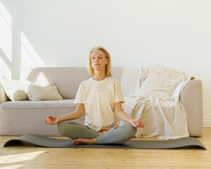Peaceful senior woman in lotus position meditation with closed eyes at home while sitting on yoga...
