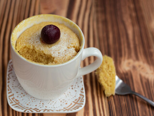homemade cupcake in a mug cooked in the microwave. Decorated with cherries and icing sugar. without one piece. Part of the biscuit on a fork lies on a wooden table. Place for text. Cherry on the cake