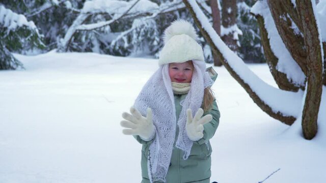 Little happy girl smiles and rejoices in winter. A child in the snow and drifts. A warm kerchief and a hat with a bumbon on a child in a winter park and forest. We meet winter. High quality 4k footage