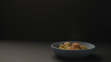 fried shrimps falling on pesto penne in blue bowl on black background
