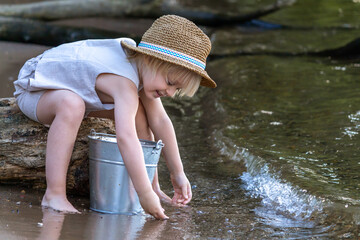 Little blond caucasian boy wearing straw hat sits on the tree log by the river and plays with metal bucket full of water. Summer kids fun theme.