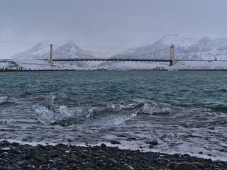 Small floating iceberg with clear surface floating on Diamond Beach near Jökulsárlón glacier lagoon, southern Iceland in winter season with suspension bridge and rugged snow-capped mountains.