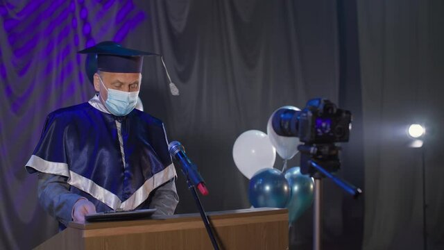 male lecturer wearing medical mask observes precautions and solemnly delivers diplomas online via video link to graduates during quarantine