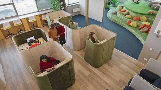 Top view shot of young people sitting in private booths in modern coworking space or start-up office and working on laptops. Men walking and discussing document