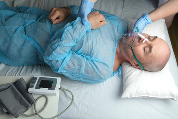 elderly man patient in blue shirt lies, the doctor puts on facial oxygen mask, concept of medical care, anesthesiology and intensive care, oxygen therapy, treatment of coronavirus, COVID-19