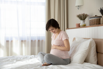 Smiling young Caucasian woman sit on bed at home bedroom touch caress big baby bump. Happy millennial female relax rest enjoy easy healthy last month of pregnancy. Parenthood, motherhood concept.