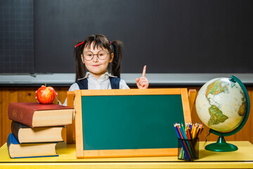 Little primary school girl in the classroom. Points his finger at the empty space for text, messages, advertising. School supplies on the desk.