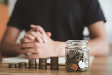 Financial saving money to plan in the future. Man wearing black shirt sitting and hands clasped with coins stack, jar, notebook on wooden desk at workspace room