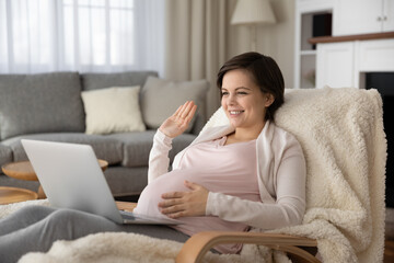 Smiling young Caucasian pregnant woman sit in armchair at home wave greet talking on video call on laptop. Happy female with pregnancy have online digital virtual communication on computer.