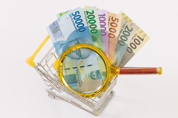 A miniature shopping cart with Indonesian currency and a magnifying glass. Economic and business concept, rising inflation and the country's economic upset. White background