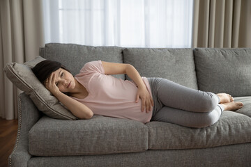 Obraz premium Unwell young Caucasian woman lying on couch touch baby bump feel unhealthy suffer from pain ache. Tired female relax on sofa touch baby bump have health problems during pregnancy. Healthcare concept.