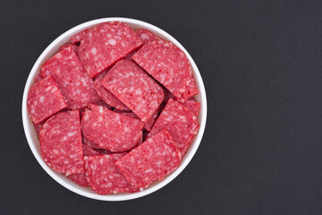 White glass bowl of sliced salami. Isolated on a dark grey background. Top view close up