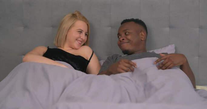 Mixed race couple lying in bed. Caucasian woman hinting at physical intimacy. African man shows readiness. Male's erection. Sexual relationship