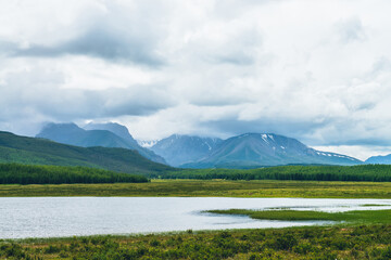 Fototapeta premium Dramatic landscape with mountain lake and forest on hills in sunlight and snowy mountains in low clouds in changeable weather. High mountains in cloudy sky and lake near green grasses and forest hills
