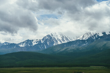 Dramatic mountains landscape with snowy mountain range among low clouds. Atmospheric highland scenery with high mountain ridge above green forest in overcast weather. Awesome view to mountain wall.