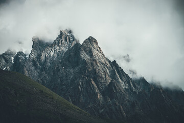 Obraz premium Dark atmospheric surreal landscape with dark rocky mountain top in low clouds in gray cloudy sky. Gray low cloud on high pinnacle. High black rock with snow in low clouds. Surrealist gloomy mountains.