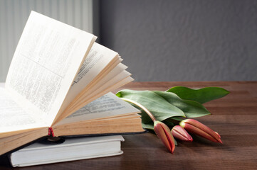 An open book on a wooden tray. Flowers on the book. A stack of books. Workplace.