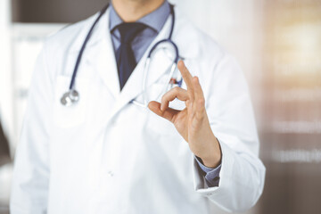 Unknown male doctor standing straight with OK sign in sunny clinic, closeup. Perfect medical service in hospital. Medicine and healthcare concept