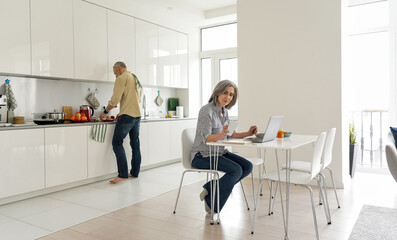 Mature mid aged wife remote working online from home office using laptop computer while senior husband cooking breakfast in kitchen helping with household work in modern kitchen apartment interior.