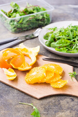 Peeled and sliced orange on a cutting board and a mix of arugula, chard and mizun on a plate on the table. Ingredients for making vitamin dietary vegetarian salad. Vertical view