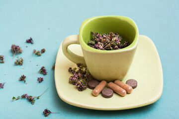 A cup with dried oregano and herbal medicinal capsules and pills on a saucer on a green background. Alternative medicine