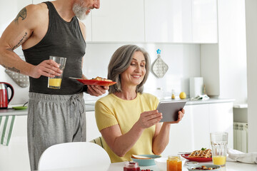 Happy older mature family couple having breakfast using digital tablet in kitchen. Smiling mid age senior husband and wife looking at pad computer enjoying morning meal together shopping online.