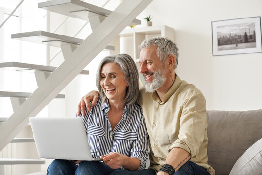 Happy Older Mid Age Family Couple Using Laptop Sit On Couch. Smiling Senior Adult Mature Man And Woman Looking At Computer Doing Ecommerce Online Shopping, Watching Tv, Having Virtual Chat At Home.