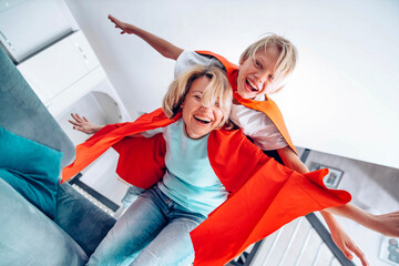 Child and mother in superhero costume playing together at home - Happy son and mom having fun in living room - Family concept - Powered by Adobe