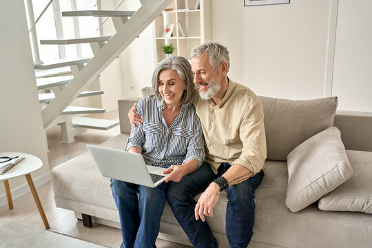 Happy old mid age family couple using laptop computer sitting on couch. Smiling senior mature husband and wife doing ecommerce online shopping, browsing web, watching tv, having video call at home.