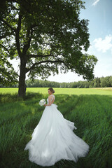 Model girl in a luxurious wedding dress with bridal flower bouquet posing in the green field near the alone tree on the summer day