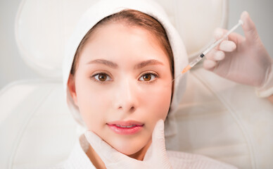 Asian woman lying on bed and cosmetologist does injections to face augmentation for anti wrinkle in...