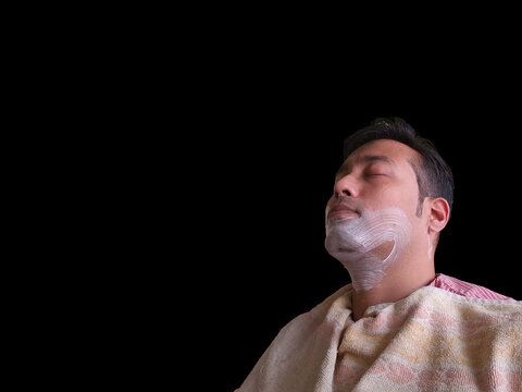 An Indian Man In Pink Shirt Wearing Yellow Towel Sitting On A Chair With Eyes Closed And Foam Applied For Shaving In Black Background. View From Side