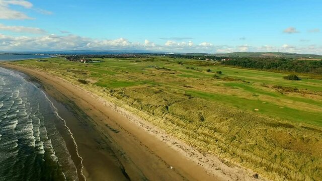 Aerial Panning The Lush Greens Of The Royal Troon Golf Club And Rugged Coast, With Blue Skies, Ocean Surf, And Exclusive Homes Nestled In The Trees