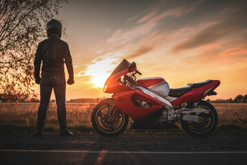 Fototapeta na wymiar A motorbiker is standing on the road in the sunset rays and looking far.