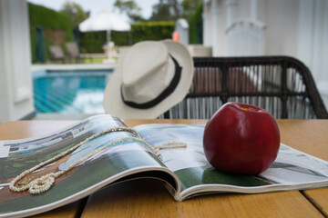 Vacation concept in villa hotel with pool. Magazine, pearl necklace and red apple on the table