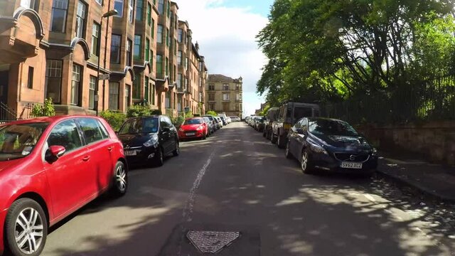 Looking Forward From A Car Down A Street As It Turns A Corner In A Historic Glasgow Neighborhood Of Victorian Townhomes, With Lush Trees, And A Bright Blue Sky