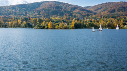 Żaglówki pływające po jeziorze, w tle góry porośnięte lasem w kolorach jesieni