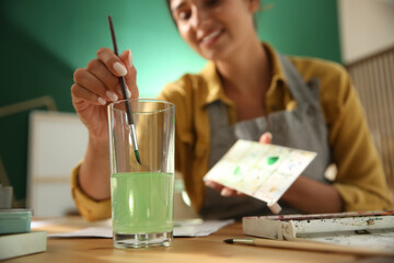 Young woman drawing at table indoors, focus on hand with brush