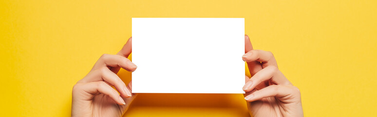 Fototapeta premium Female hands hold a blank sheet of paper on a yellow background. Advertising space