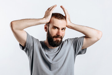 Portrait of handsome young man frowning while holding hands near head on white background with space for advertising mock up