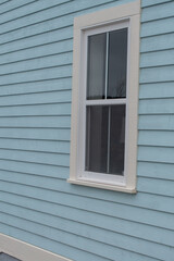 The exterior wall of a baby blue country style house with narrow wood cape cod siding. There are two small double hung windows with white trim. Lace curtains are hanging in the closed glass windows.